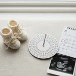 A physical OB wheel calculator is laid on a clean, light-colored surface, next to a pair of small knitted baby booties and a calendar with a heart on a future date.
